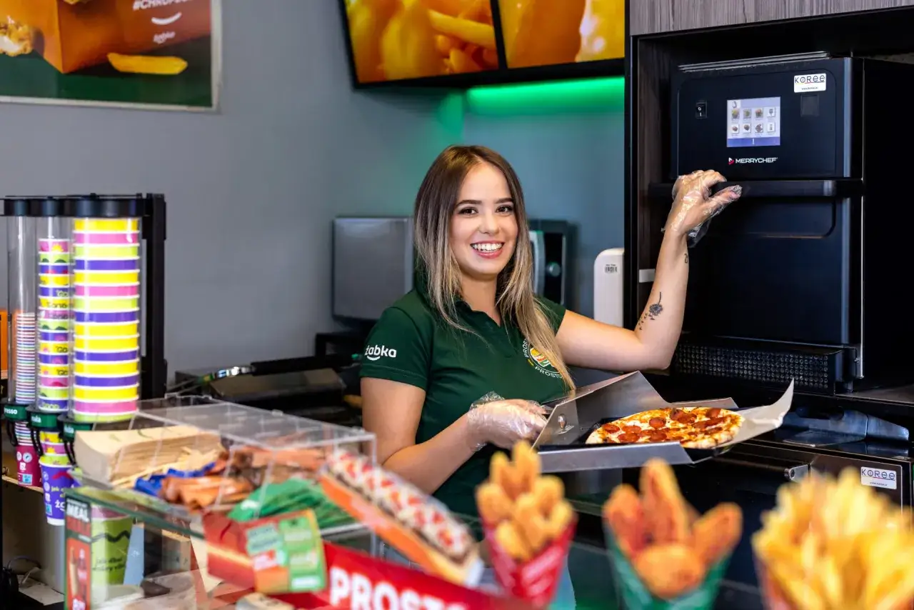 Uśmiechnięta pracownica Żabki przygotowuje pizzę. Zastanawiasz się, ile zarabia się w Żabce?