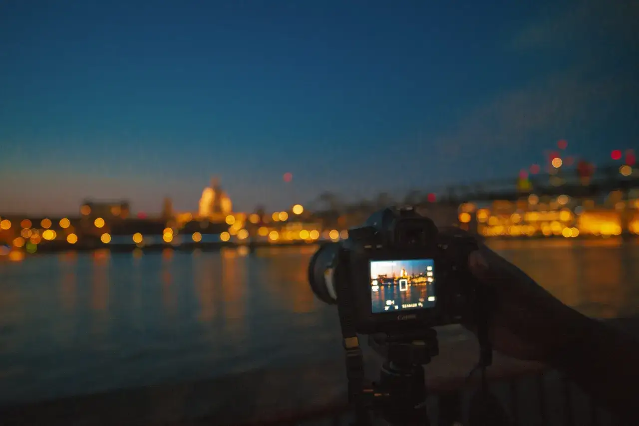 Une main tient un Canon EOS 2000D pour une photo de nuit, capturant des lumières de ville floues sur l'eau.