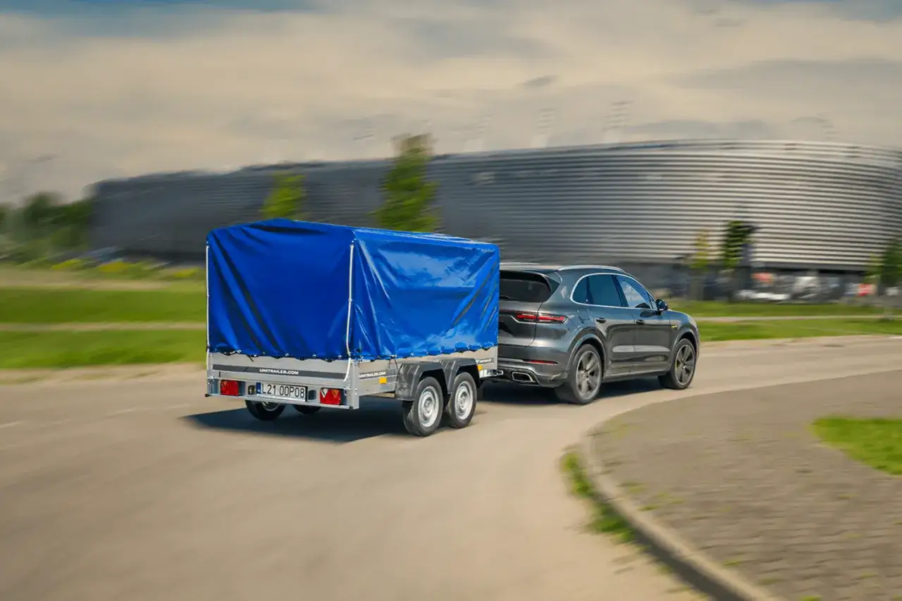 Szary Porsche Cayenne z przyczepą pod niebieską plandeką jedzie drogą obok nowoczesnego budynku.