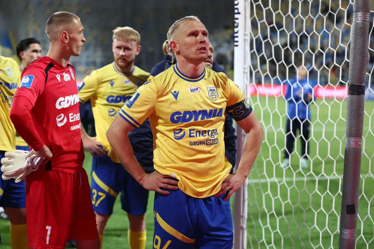 Piłkarze Arki Gdynia w żółtych strojach, jeden z nich to trener Arki Gdynia, stoją przy bramce.
