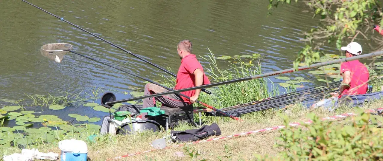 Dwóch wędkarzy siedzi nad wodą, jeden z nich właśnie wyciąga rybę z sieci. W tle widać spokojny **zbiornik zaporowy** z liliami wodnymi.