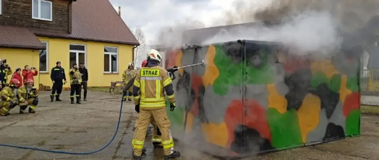 Strażak używa lancy do gaszenia pożaru na poligonie. W tle widać budynek i innych strażaków.