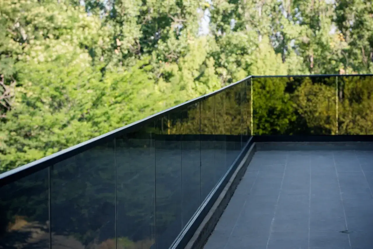 Barandilla de terraza con vidrio laminar de seguridad oscuro, reflejando &aacute;rboles verdes.