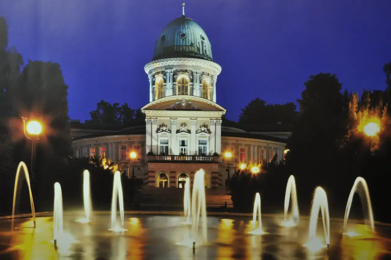 Nocny widok na zabytkowy budynek w Lądku Zdroju z fontanną w podświetlonym basenie.