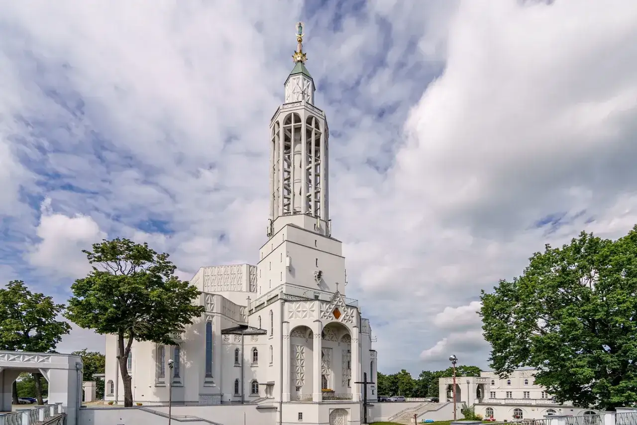 Biały, nowoczesny kościół św. Rocha z charakterystyczną, ażurową wieżą góruje nad zielenią drzew.