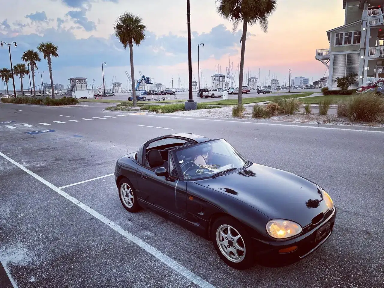 Czarny Suzuki Cappuccino z otwartym dachem na tle portu jachtowego o zachodzie słońca.