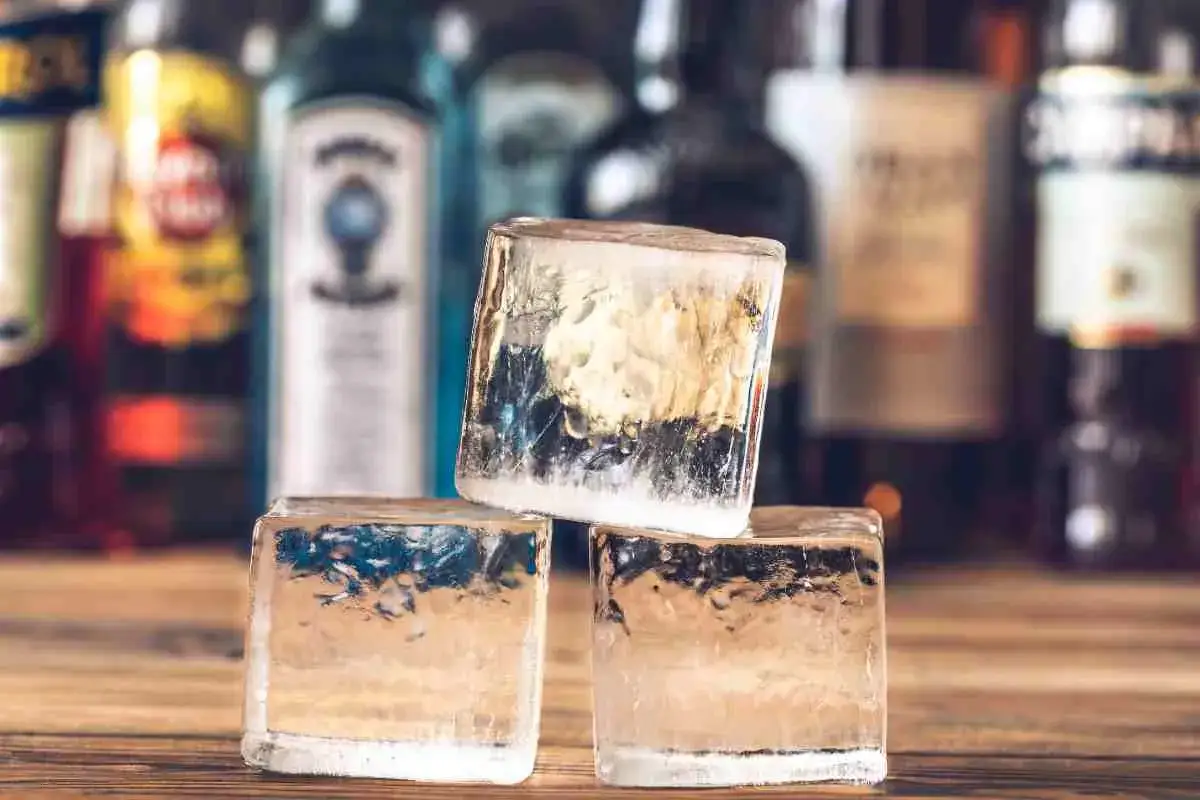 Tre cubetti di ghiaccio impilati su un tavolo di legno, con bottiglie di liquori sfocate sullo sfondo. Perfetti per un drink rinfrescante.