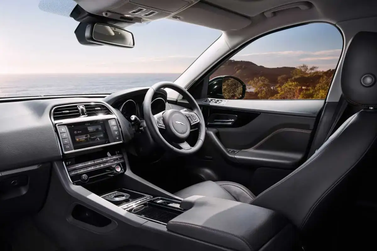 Interior de un coche de lujo, mostrando el lado derecho con el volante, pantalla táctil y asientos de cuero. El paisaje exterior es un mar y colinas.