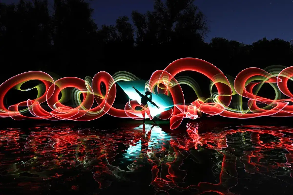 Una figura danzante crea un vortice di luce rossa e gialla con la tecnica del light painting, riflessa sull'acqua scura.