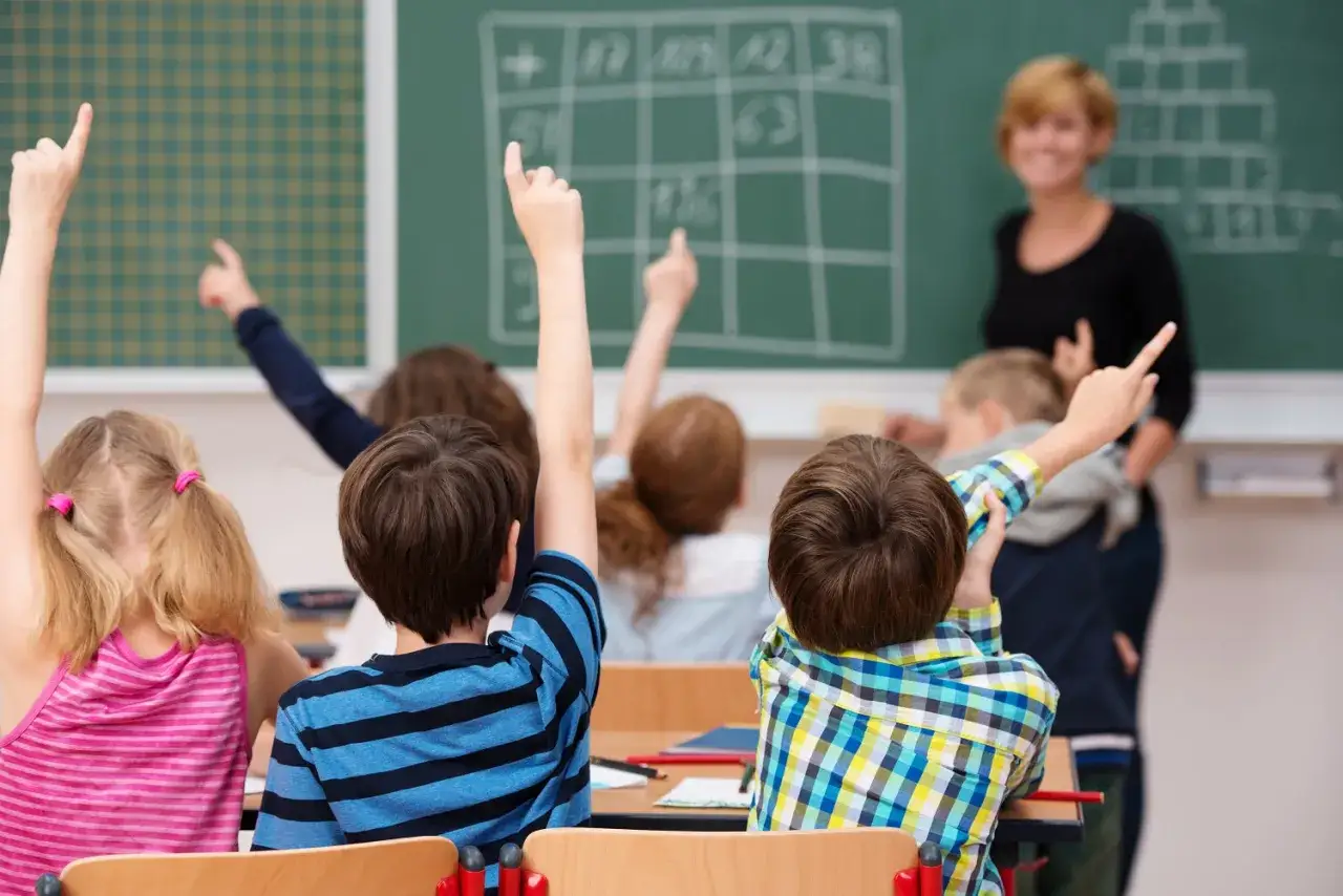 Viele Kinder in der Schule strecken die Hände hoch, um eine Frage zu beantworten. Die Lehrerin lächelt.