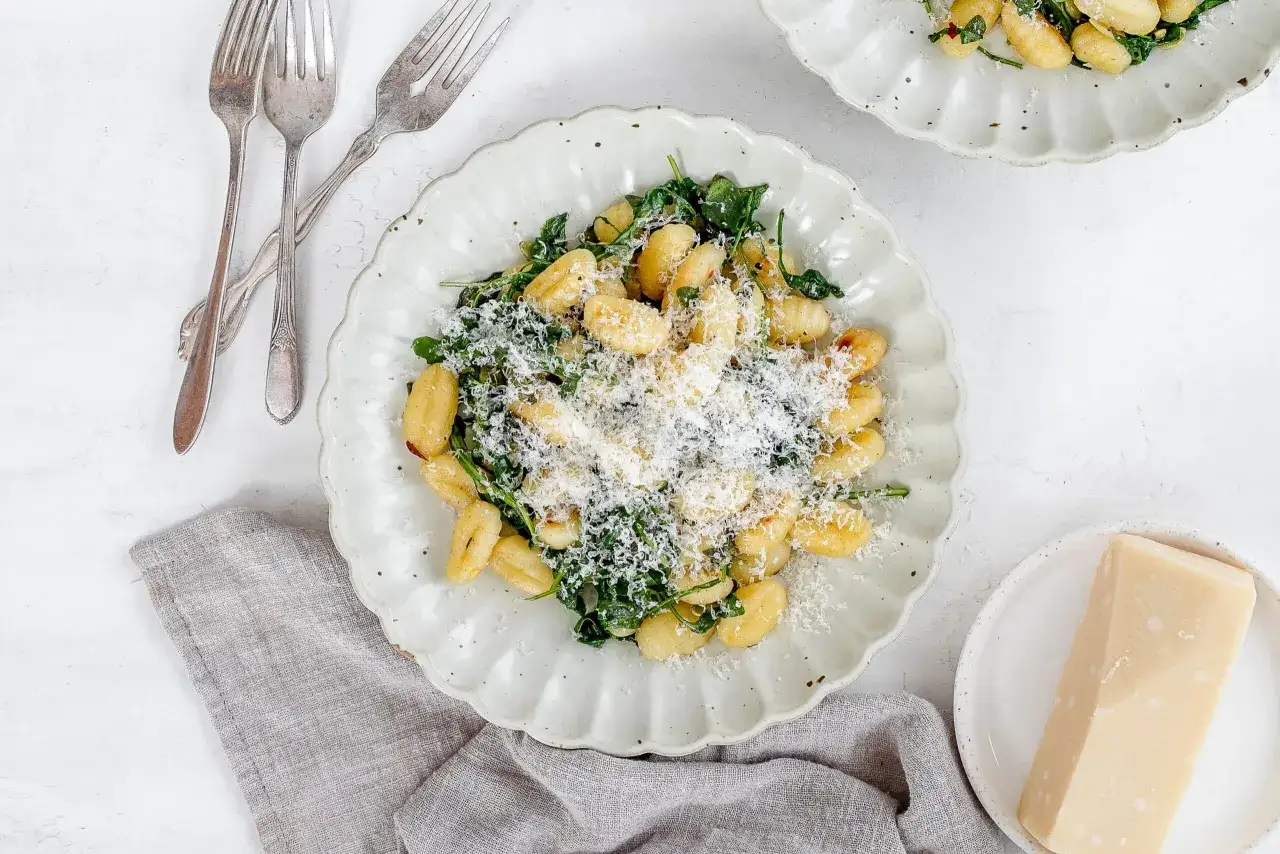 En tallrik med gyllene, stekta gnocchi med gröna blad och riven parmesan. En bit parmesan och bestick ligger bredvid.