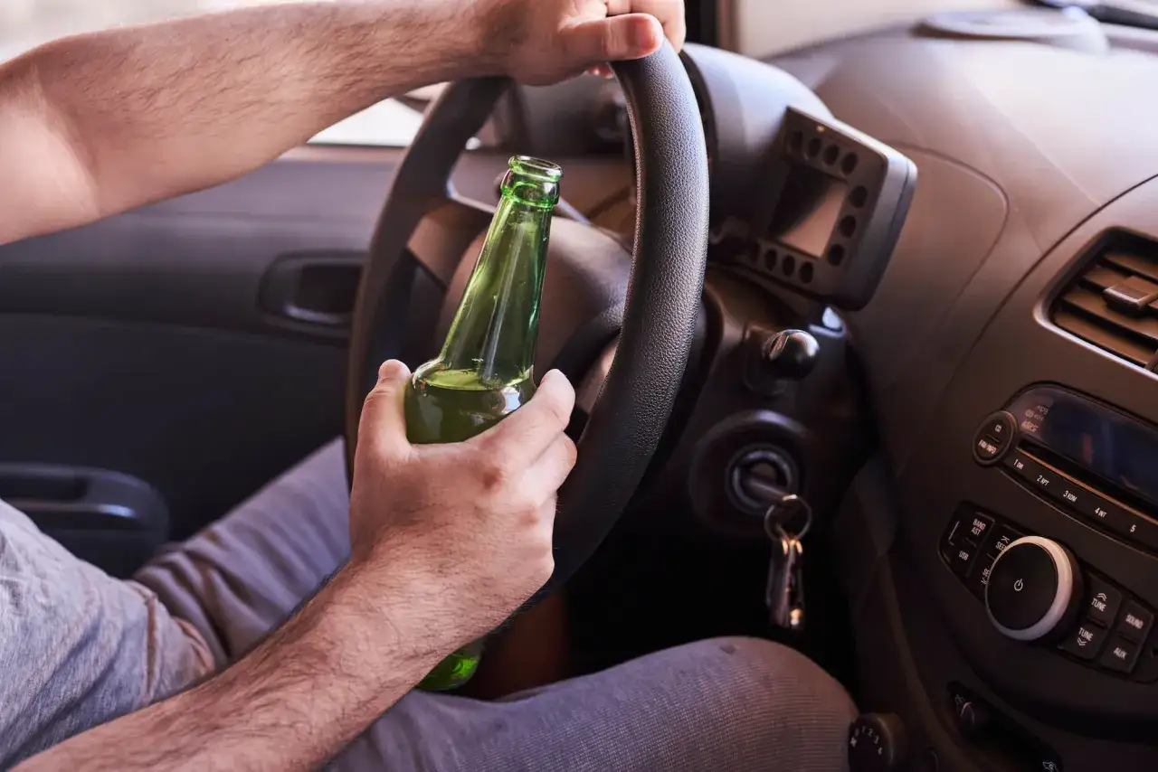 Kierowca trzyma butelkę piwa za kierownicą. Prowadzenie pojazdu pod wpływem alkoholu jest nielegalne i niebezpieczne, nie ma dopuszczalnej ilości alkoholu.