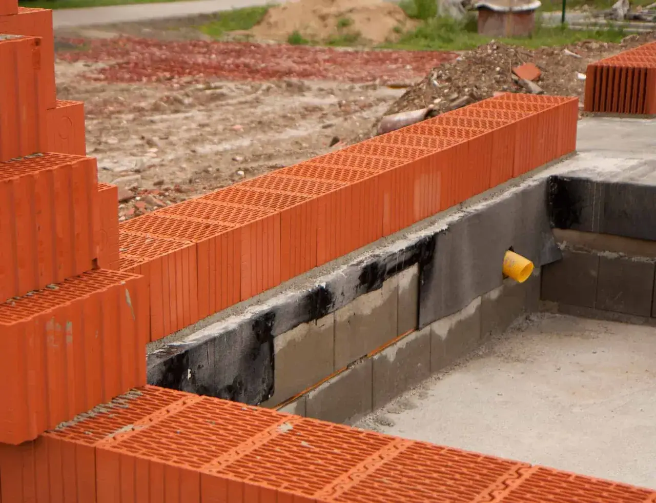 Postępujące murowanie ścian z porothermu na fundamencie. Widać ułożone pustaki ceramiczne i fragmenty izolacji.