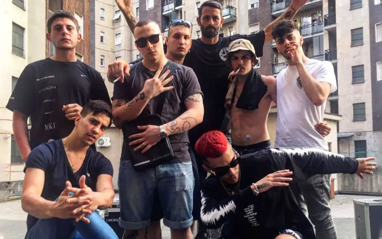 Un gruppo di ragazzi, forse la dark polo gang, posa con atteggiamento sicuro. Alcuni fumano, altri mostrano tatuaggi.