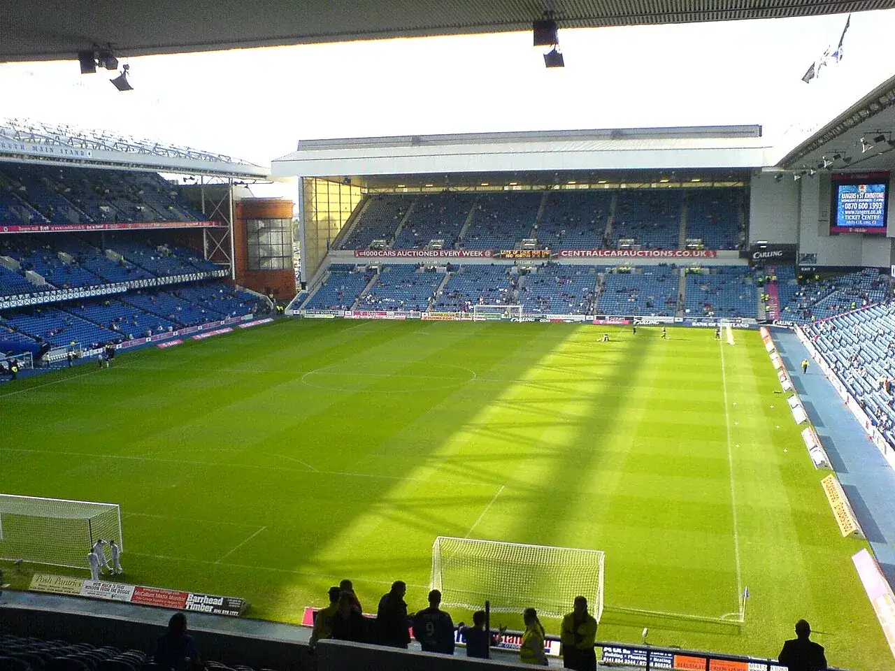 Piłkarze z Glasgow na zielonej murawie stadionu, gotowi do gry. Słońce rzuca długie cienie na boisko.