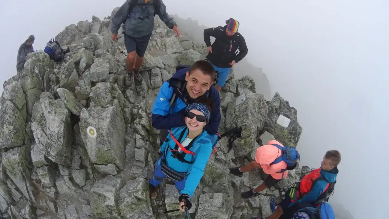 Grupa turystów na skalistym szczycie, otoczona mgłą. Jedna z kobiet, mała wysoka, robi sobie selfie z partnerem.