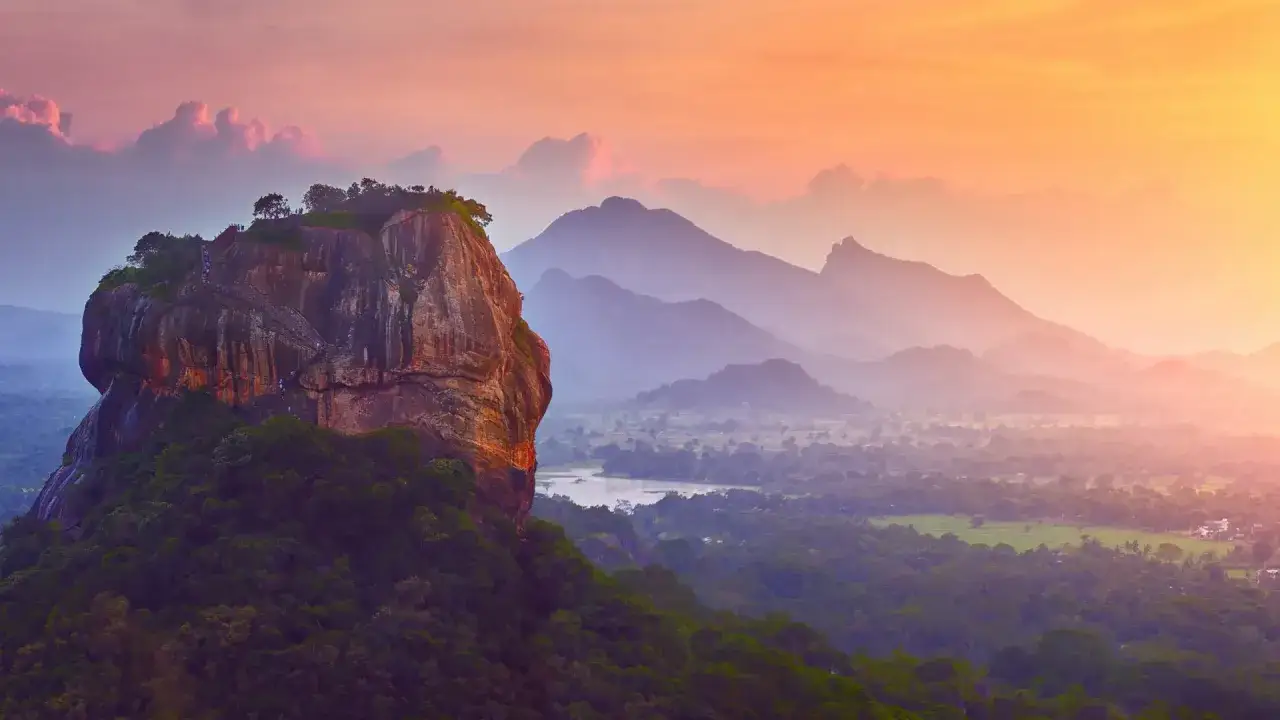 Solnedg&aring;ng &ouml;ver Sri Lanka. En imponerande klippa, likt Adam's Peak, reser sig ur frodig gr&ouml;nska. M&aring;nga berg i bakgrunden badar i gyllene ljus.