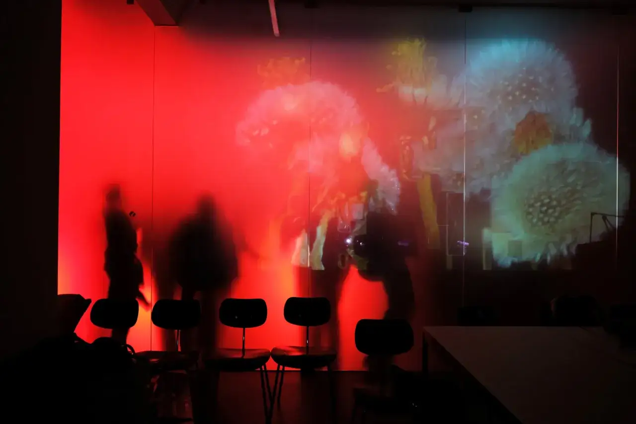 Silhouetten von Menschen vor einer roten Wand mit Projektionen von Pusteblumen. Einblicke in die berlin design week.