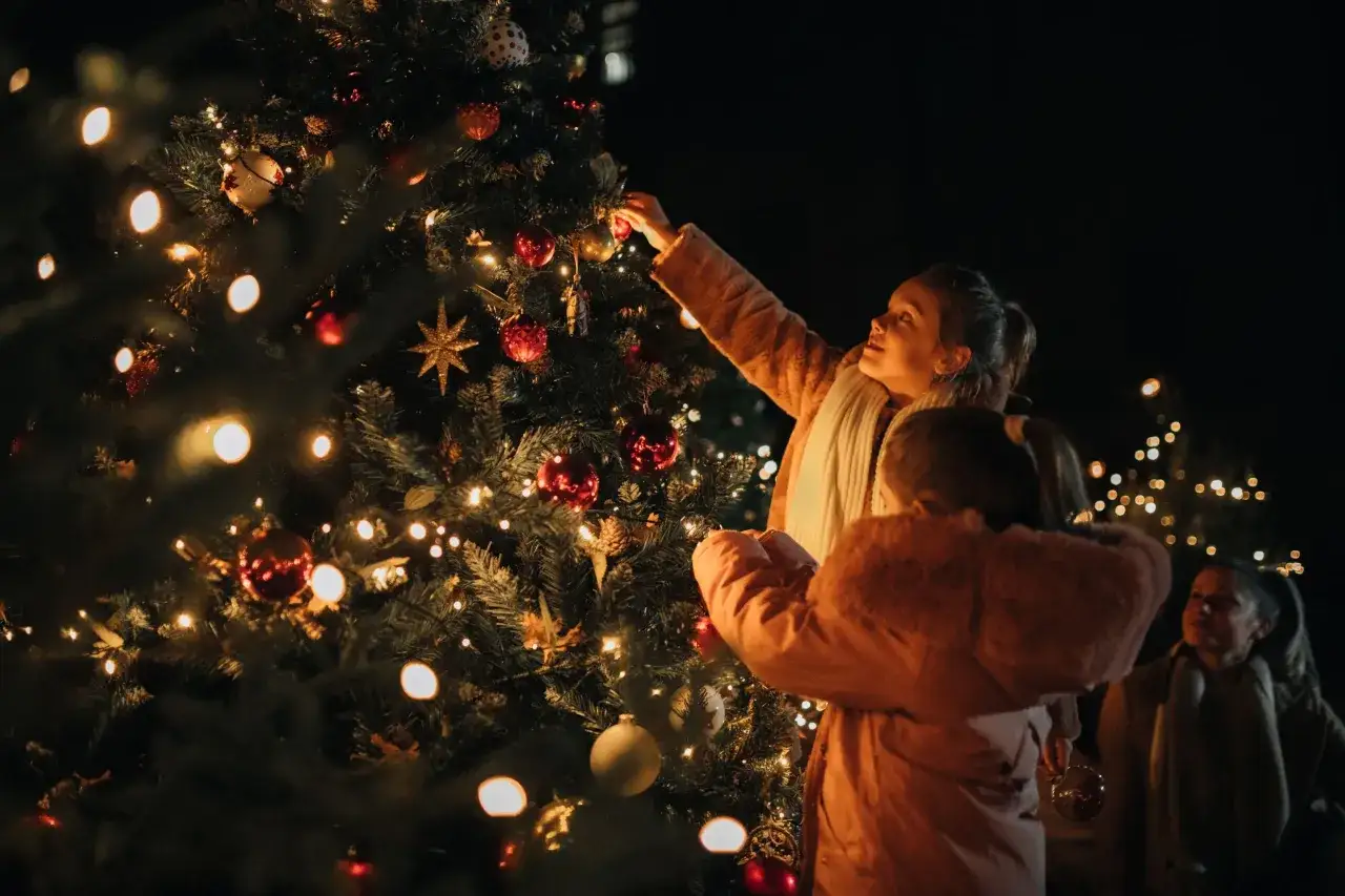 Bambini decorano un albero di Natale illuminato, creando un'atmosfera magica. Le luci scintillanti e le palline rosse sembrano sussurrare le parole del Natale.