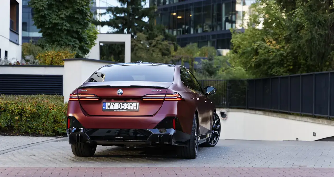 Eleganckie BMW 550e w kolorze bordowym prezentuje się z tyłu na tle nowoczesnej architektury i zieleni.