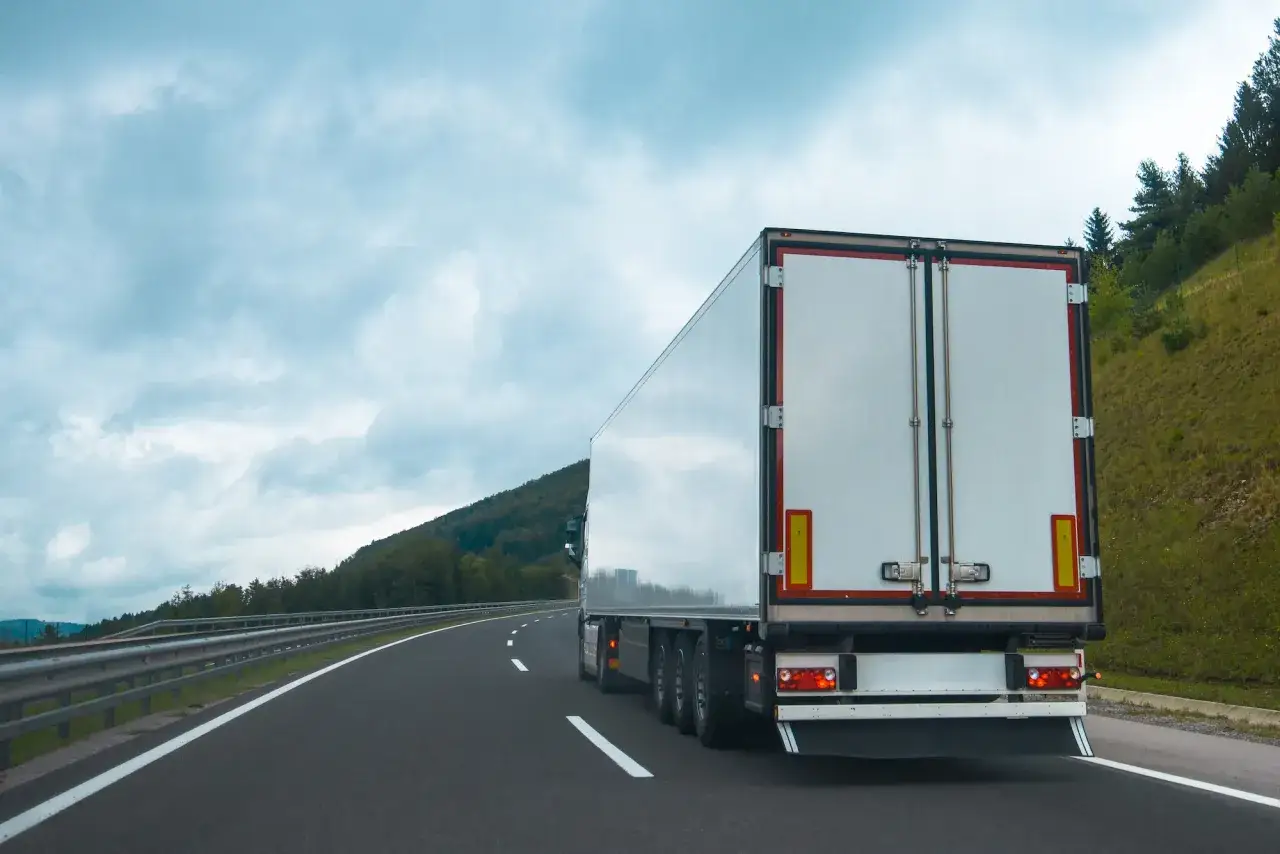 Ciężarówka na autostradzie, z tyłu widoczne białe drzwi naczepy. Na poboczu zielone drzewa i pagórki. Tablice wyróżniające przepisy są częścią oznakowania drogowego.