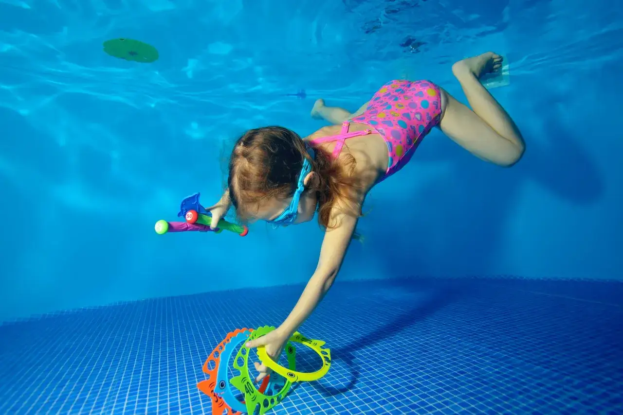 Bambina con occhialini e costume a cuoricini esplora il fondo della piscina, raccogliendo giochi. Un modo divertente per imparare a nuotare!