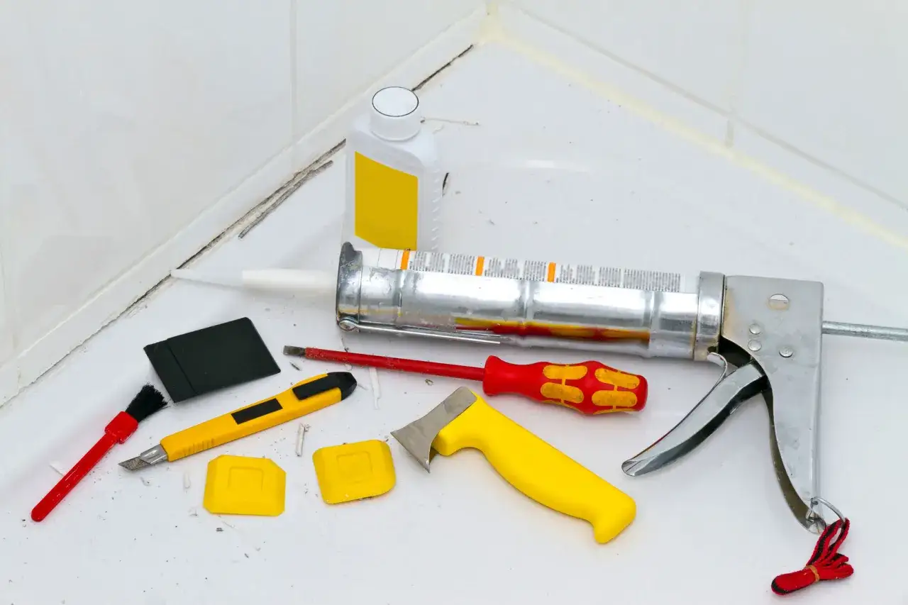 Werkzeuge zum silikon von fliesen entfernen: Kartuschenpresse, Cuttermesser, Spachtel, Bürste und eine Flasche Reiniger.