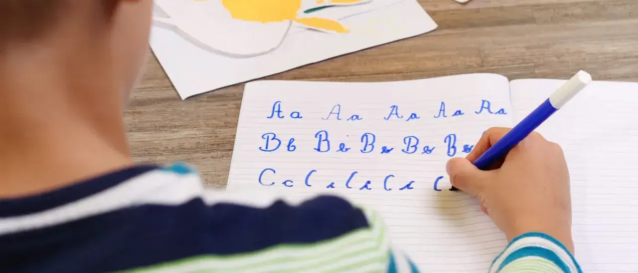 Niño practicando grafomotricidad de letras cursivas en un cuaderno. Se ven las letras A, B y C, y un rotulador azul.