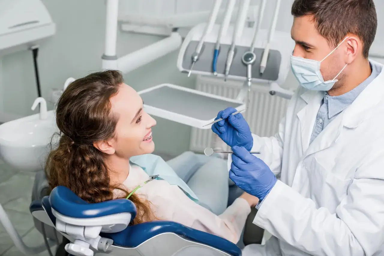Dentysta w masce bada uśmiechniętą pacjentkę z aparatem ortodontycznym.