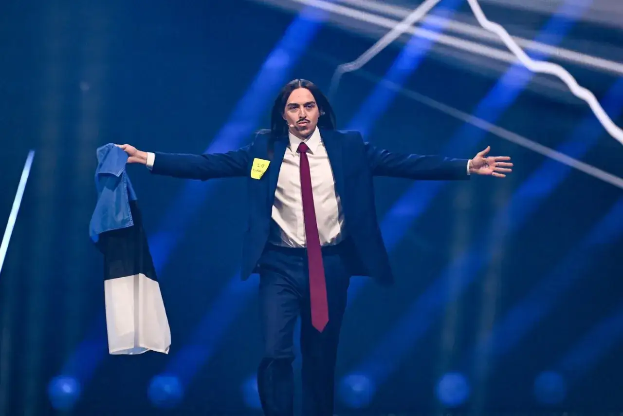 Tommy Cash w garniturze i krawacie, trzymający flagę Estonii, na tle niebieskich świateł.