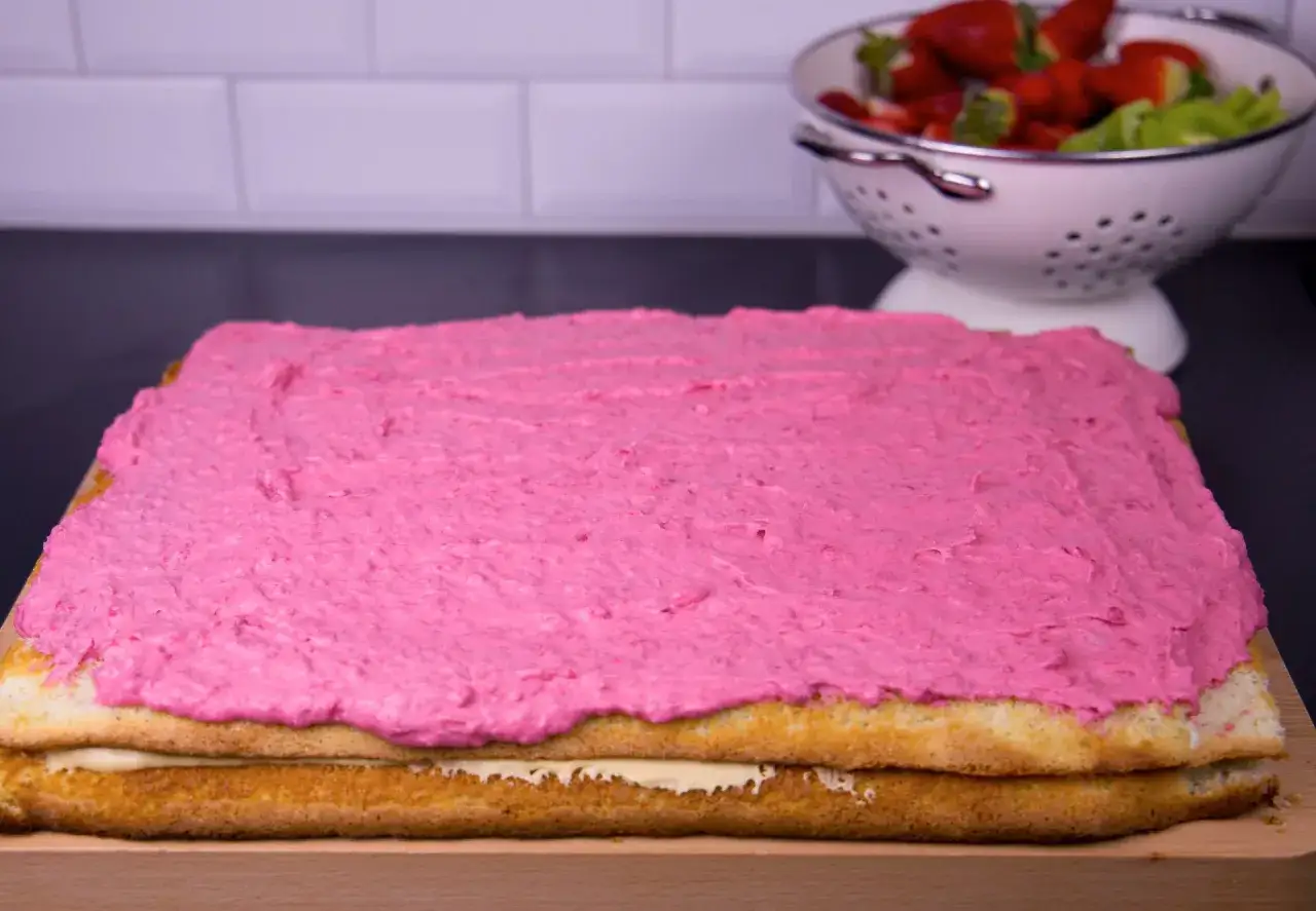 En t&aring;rtbotten i l&aring;ngpanna med rosa glasyr och en kr&auml;mig fyllning. Bredvid st&aring;r en sk&aring;l med f&auml;rska jordgubbar och kiwi.