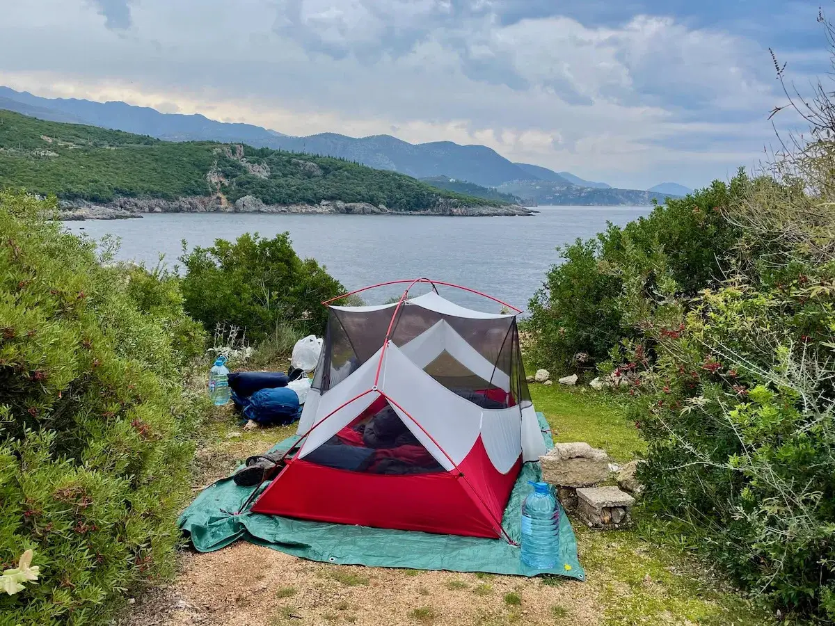 Tienda de campaña roja y blanca junto al mar. El paisaje montañoso y la vegetación exuberante sugieren que viajar a Albania es peligroso, pero la vista es impresionante.
