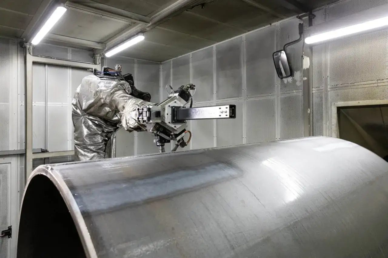 Robot w srebrnym kombinezonie wykonuje precyzyjne szlifowanie metalowej rury w hali produkcyjnej.