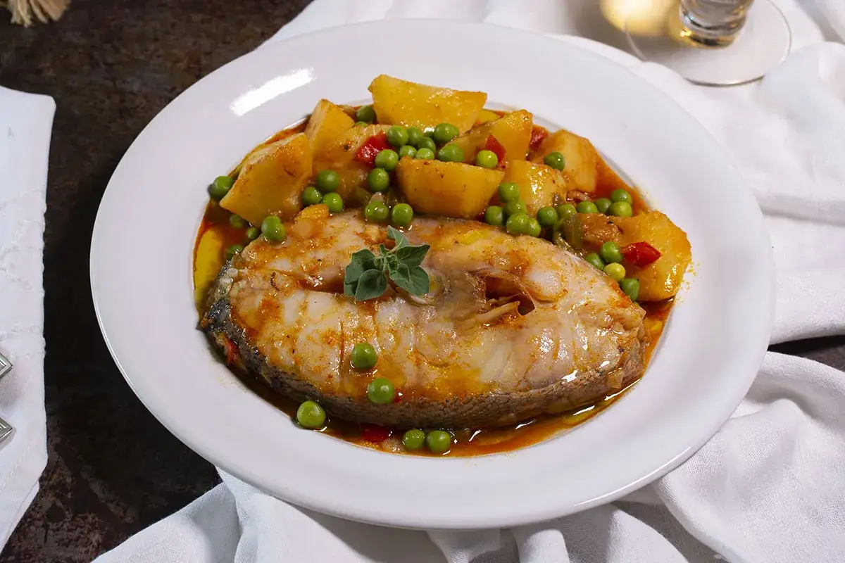Un generoso filete de abadejo pescado en una salsa rojiza con patatas y guisantes, adornado con una ramita de orégano.