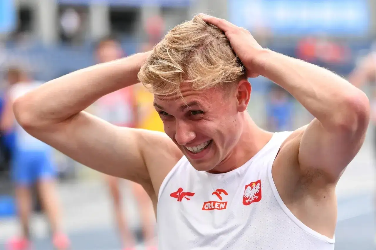 Filip Ostrowski, lekkoatleta, z uśmiechem na twarzy i rękami na głowie, w białej koszulce z orłem.