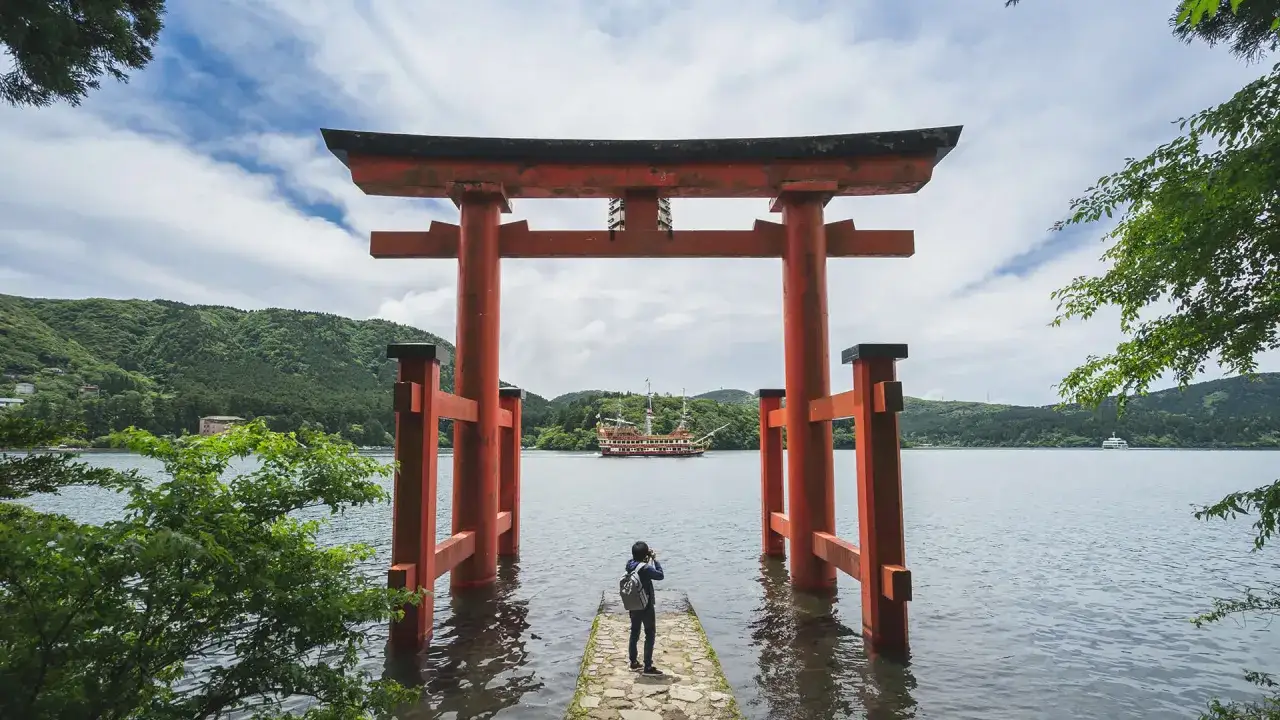 Ein Torii-Tor ragt aus dem Wasser, dahinter ein Schiff auf einem See. Ein Reisender fotografiert die Szene – ein Traum von Work and Travel Japan.