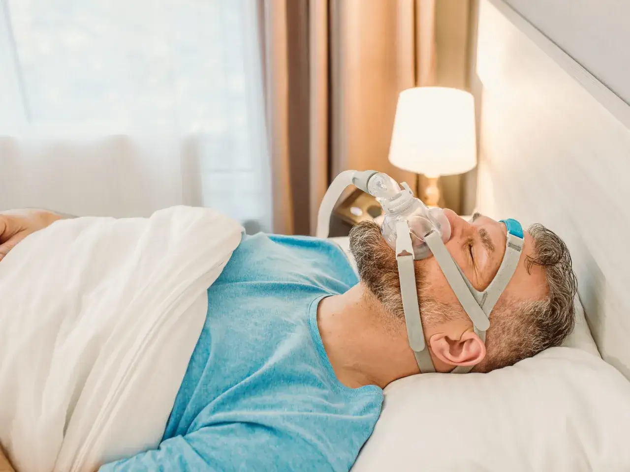 Mann mit Schlafapnoe-Maske im Bett. Die Maske hilft ihm, nachts frei zu atmen.