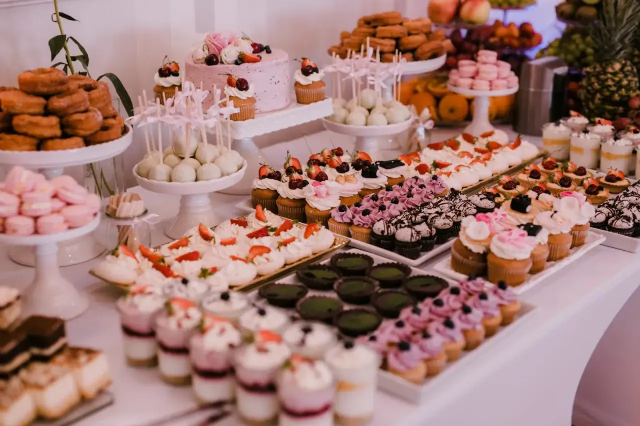 Bogactwo słodkości na weselne przyjęcie: tort, babeczki, makaroniki, pączki i deserki w pucharkach.