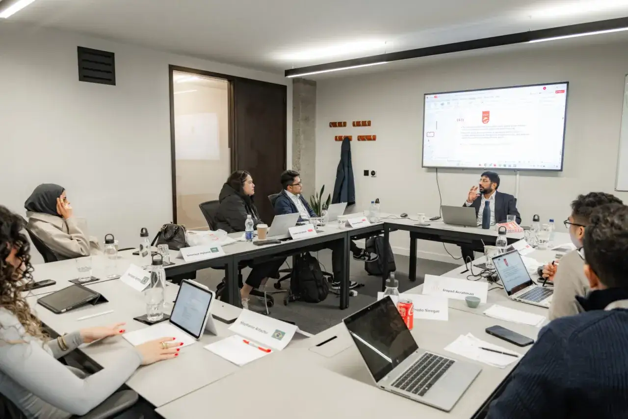 Participants in a CEO training program engage in a discussion, with a presenter at the front and attendees at a conference table with laptops.