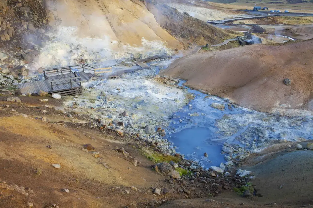 Krajobraz Gunnuhver hot springs z drewnianymi kładkami prowadzącymi przez parujące, błękitne źr&oacute;dła i skaliste zbocza.