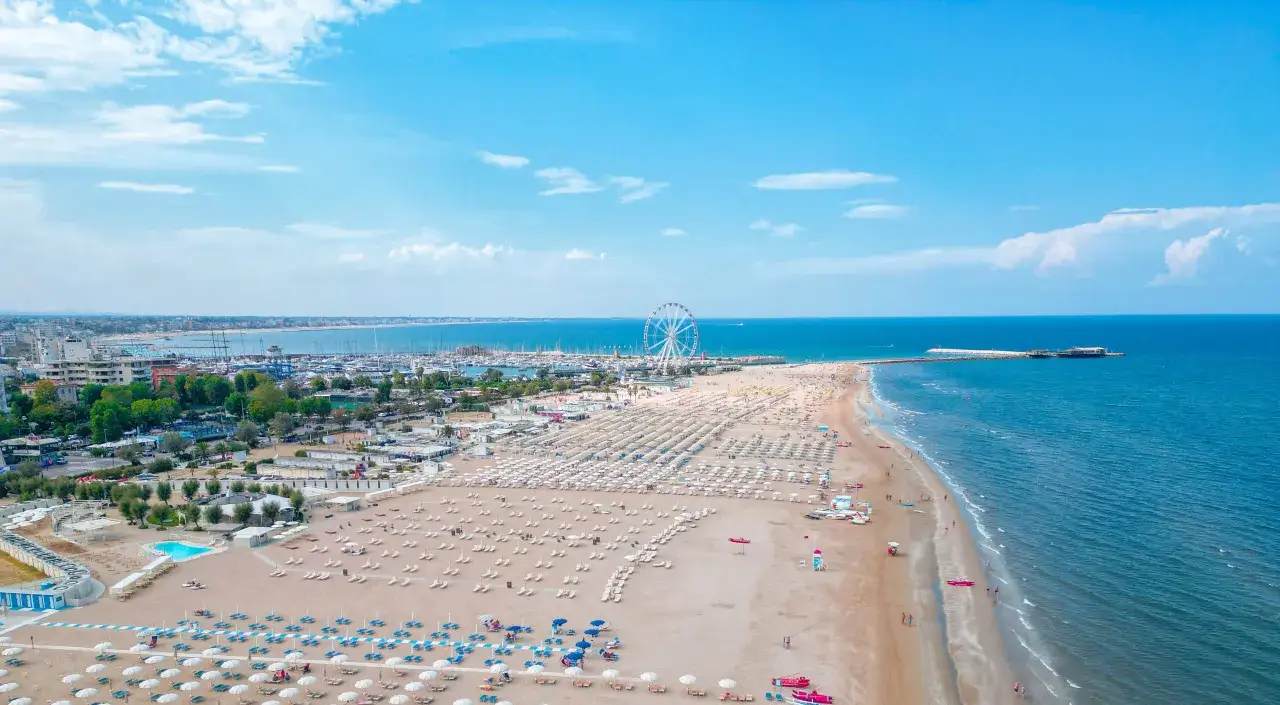 Vista aerea di una spiaggia affollata con ombrelloni e lettini, un porto turistico e la ruota panoramica, tipici dei quartieri di Rimini.
