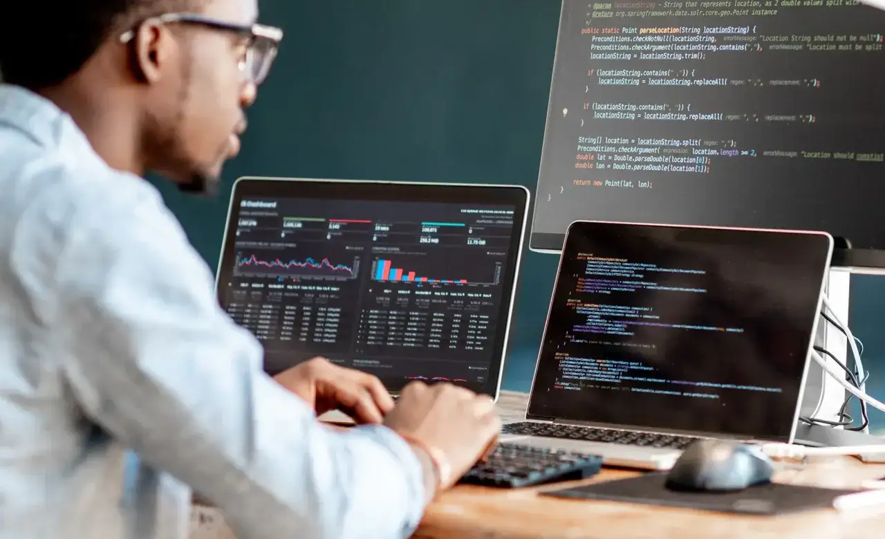 Mann mit Brille arbeitet an Laptops, die Code und Daten anzeigen. Jobs im Home Office sind flexibel und produktiv.