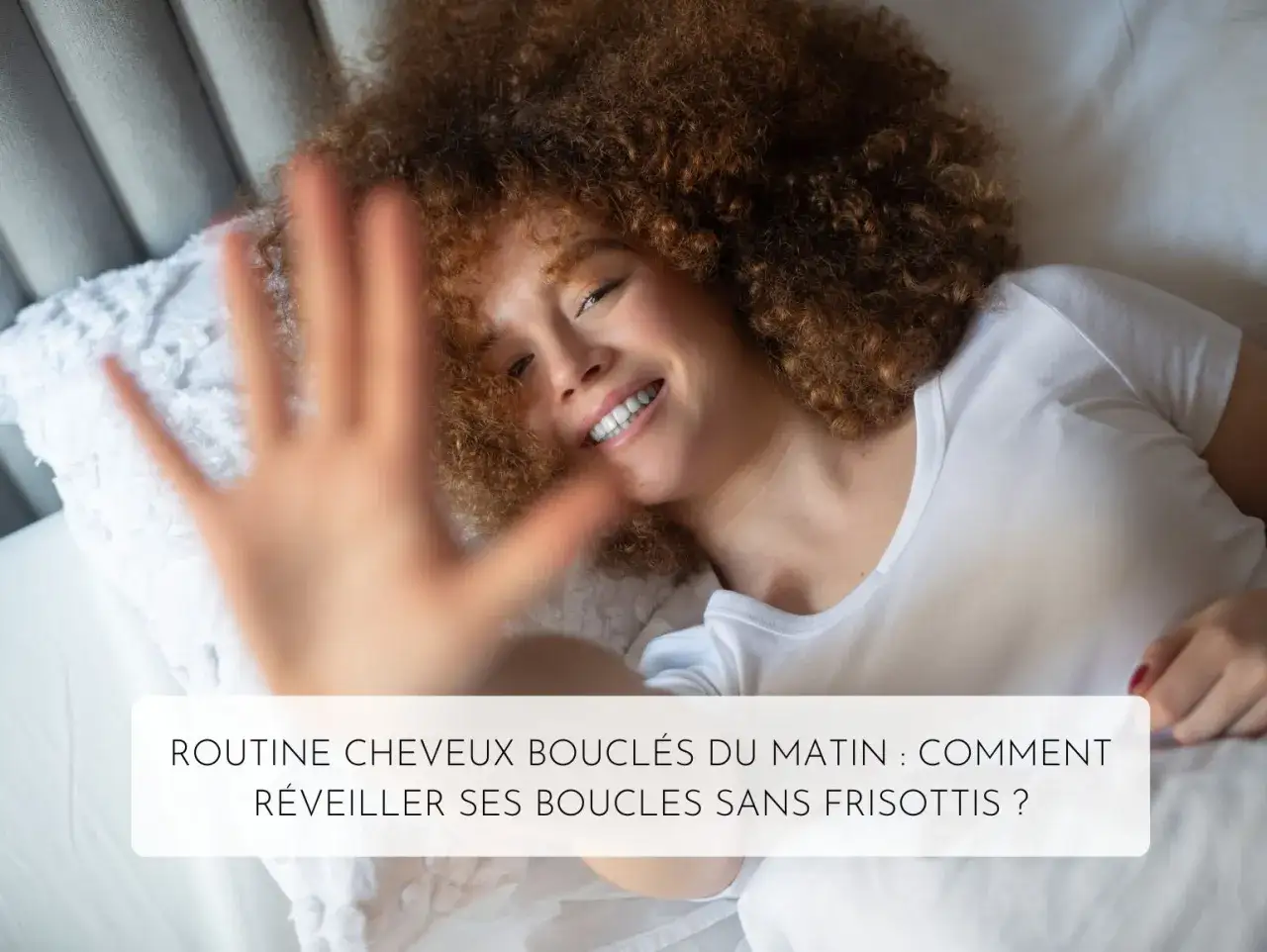 Routine matinale pour cheveux bouclés : comment réveiller ses boucles sans frisottis ? Une femme souriante aux cheveux roux et bouclés se réveille dans un lit.