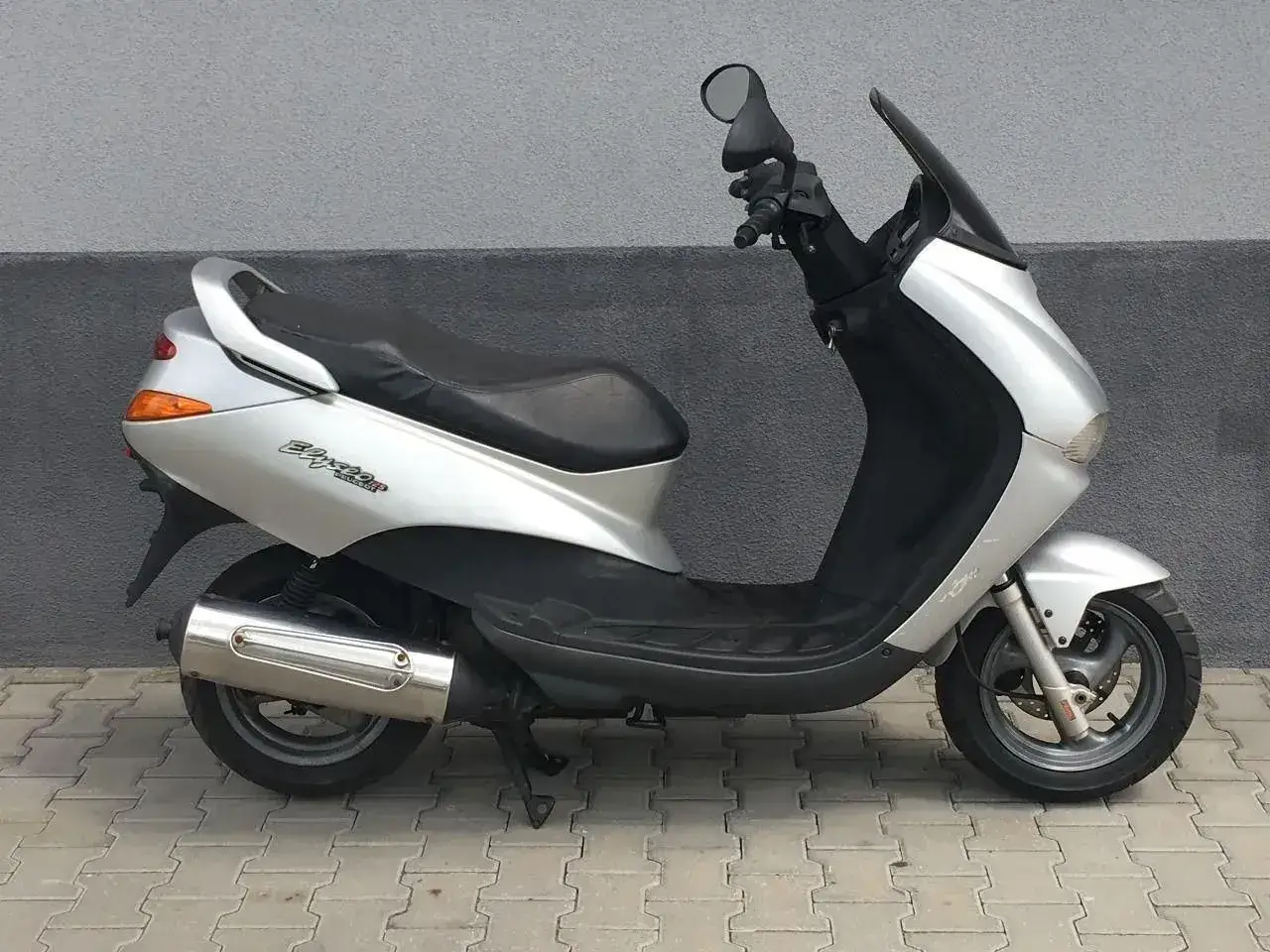 Srebrny skuter Peugeot Elyseo 125 z czarnym siedzeniem i przyciemnianą szybą, zaparkowany na chodniku.