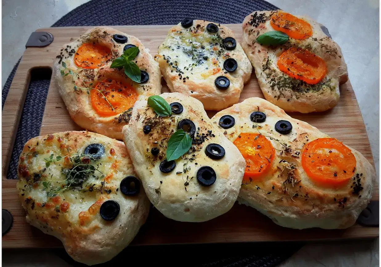 Mini ciabatty z oliwkami, pomidorkami i ziołami, ułożone na desce.