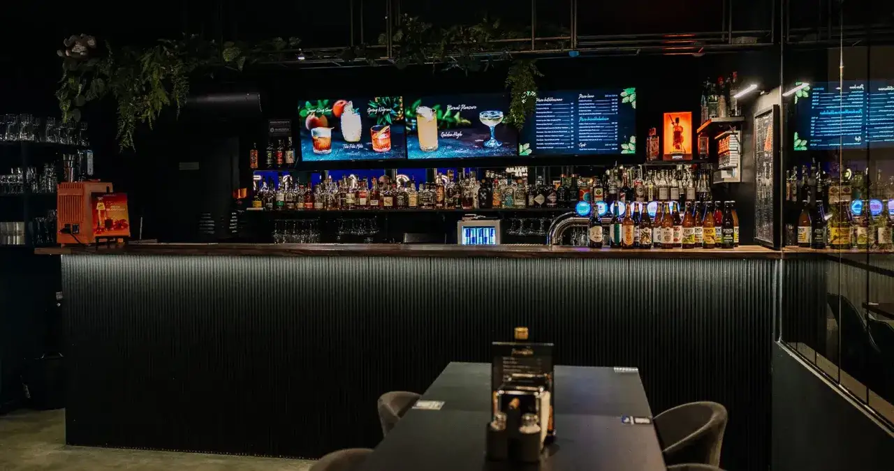 Nowoczesny bar z podświetlanym blatem, półkami pełnymi alkoholi i ekranami z drinkami. Idealne miejsce na wieczór kawalerski.