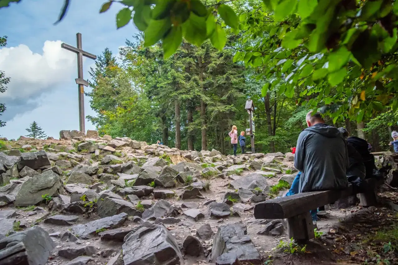 Kamienisty szczyt z drewnianym krzyżem, gdzie znajduje się najniższa góra w Polsce. Ludzie odpoczywają na ławkach, podziwiając widok.