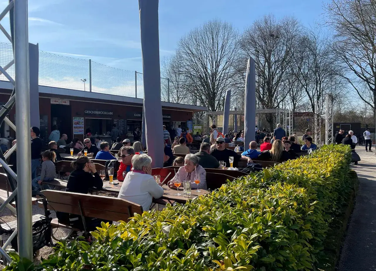 Menschen genie&szlig;en das sonnige Wetter in Carles Biergarten. G&auml;ste sitzen an Holztischen, trinken und unterhalten sich.