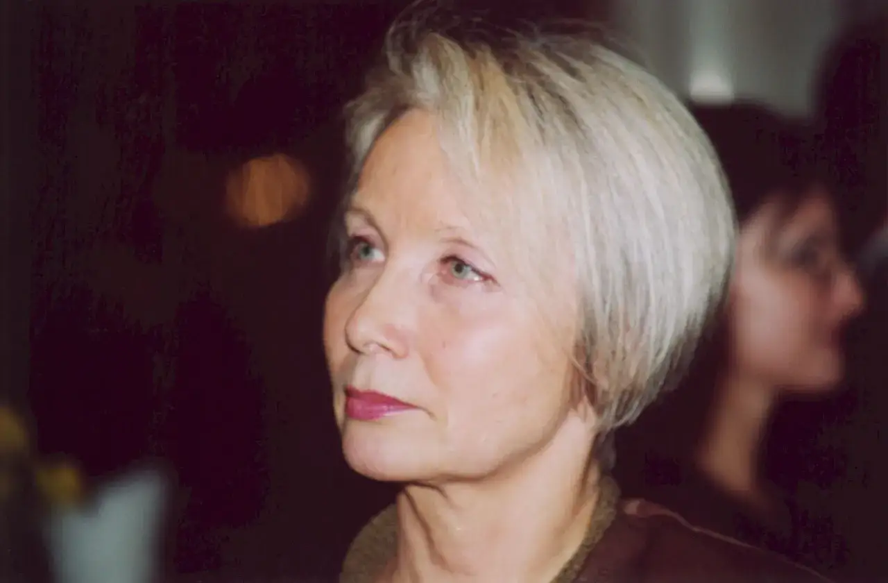 Portret kobiety z siwymi włosami, patrzącej w bok. W tle niewyraźna postać. Dziś pola raksa.