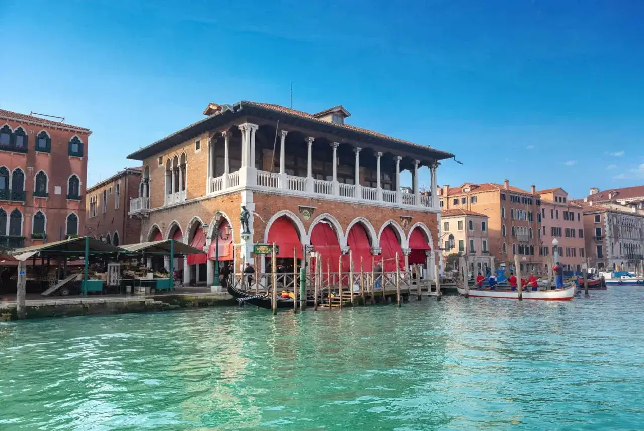 Le march&eacute; du Rialto &agrave; Venise, avec ses arcades rouges et ses gondoles, s'anime sous un ciel bleu.